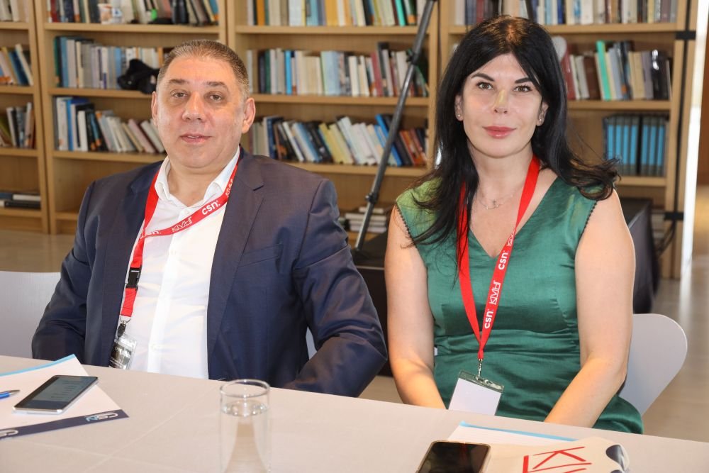Andrii Dukhno and Olga Khvorost seated side by side at a conference table, red lanyards around their necks, in front of bookshelves.